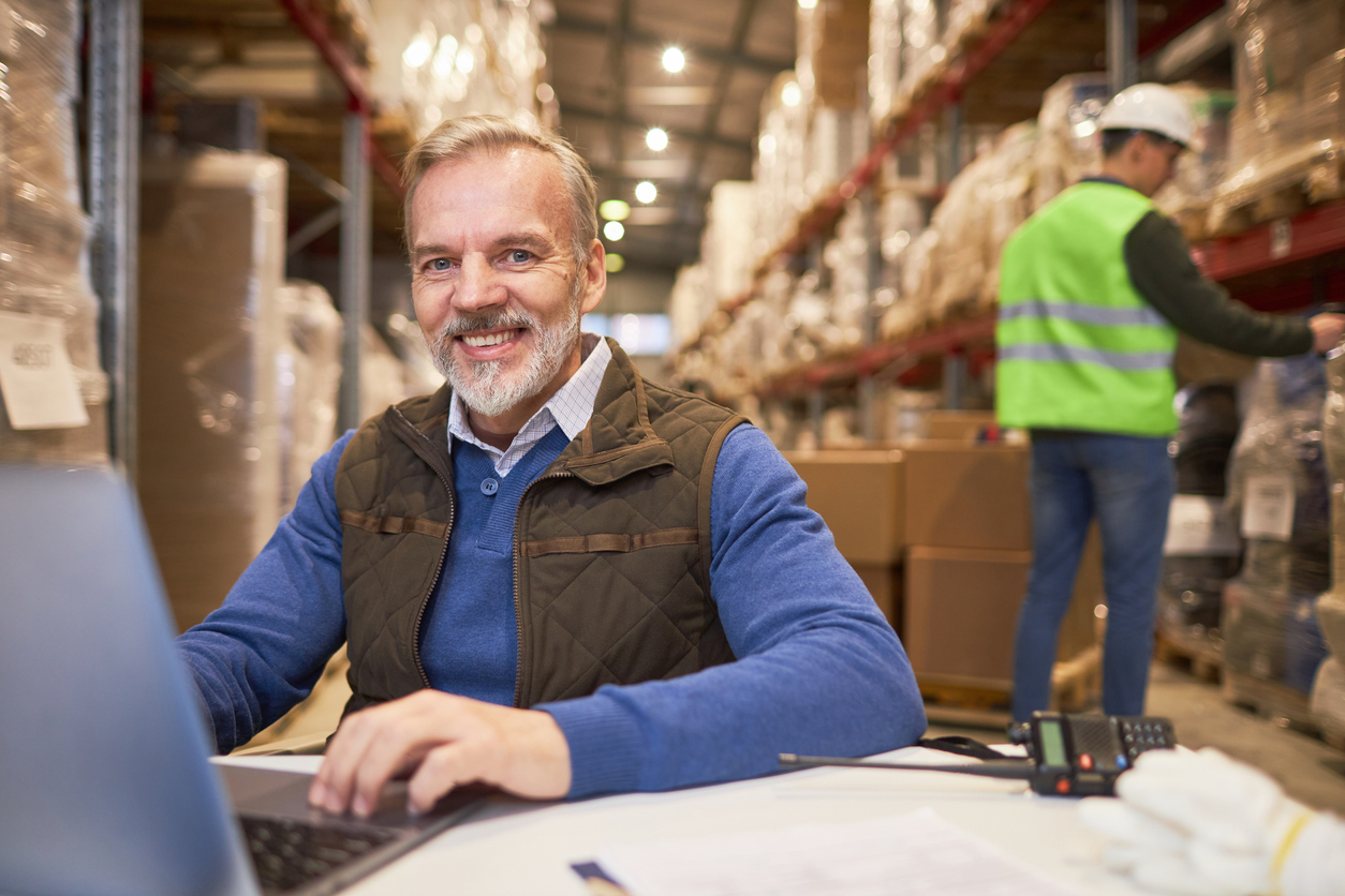 Portrait of smiling senior man using laptop in warehouse while managing distribution business