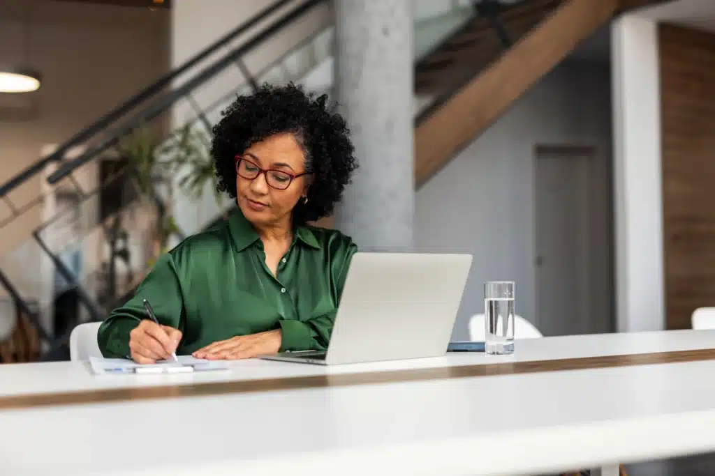 A professional woman in a modern workspace writes in a notebook while using her laptop, demonstrating focus and productivity; the setting complements professional undertakings and goal-oriented tasks.