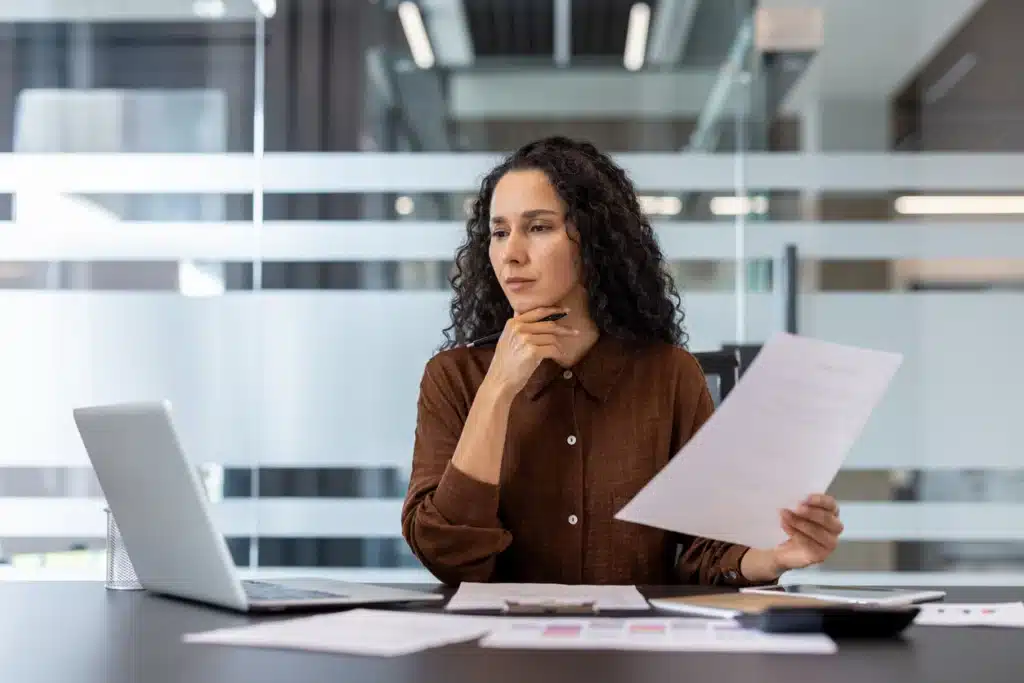 A focused businesswoman is reviewing documents and working on a laptop in a modern office setting. She appears thoughtful.