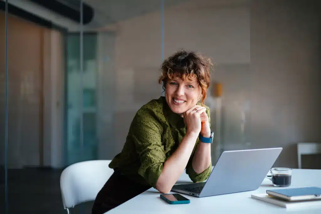 A cheerful woman sits at a white desk in a contemporary office, leaning on her hands beside a laptop, coffee, and notebook. The scene conveys productivity, focus, and a positive work atmosphere.
