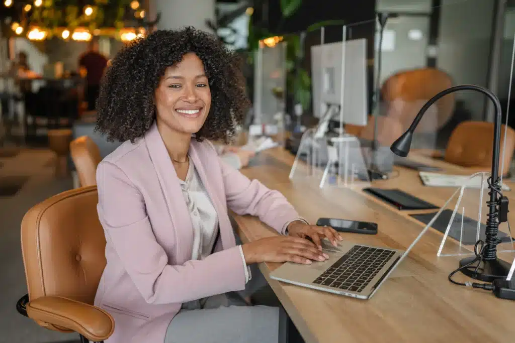 Smiling black business woman using a laptop in a contemporary co working space, working remotely and looking at the camera, symbolizing professionalism and career success